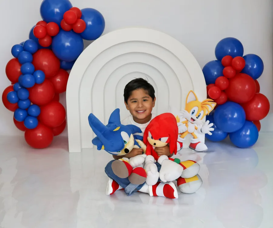 Smiling boy hugging Sonic and Knuckles plushies in front of a white rainbow arch and red and blue balloon garlands